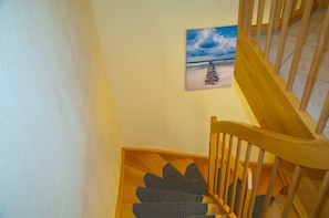 Una escalera de madera con un felpudo azul en la parte inferior, una pared con un cuadro de una escena de playa y una pared blanca a la izquierda.