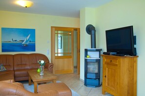A living room with a brown leather sofa, a wooden coffee table, a painting of a sailboat, a wood-burning stove, and a television on a wooden cabinet.