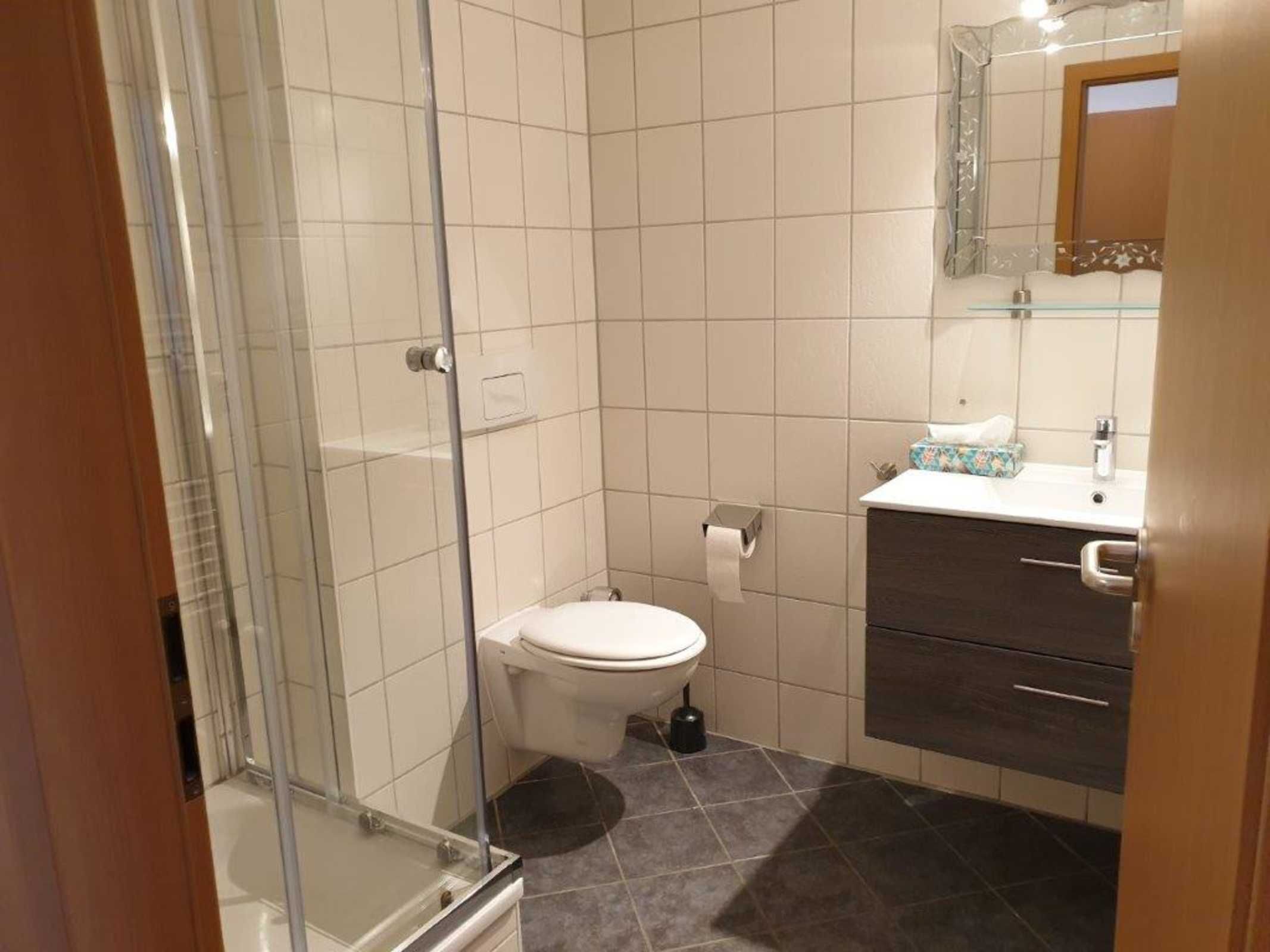 Ein Badezimmer mit Duschkabine aus Glas, Toilette, Waschbecken mit Spiegel darüber und einem dunklen Schrank.