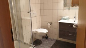 Um banheiro com box de vidro, vaso sanitário, pia com espelho e armário de cor escura.