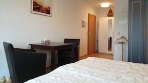 Quarto de hotel com cama, escrivaninha, duas cadeiras, cabideiro e porta para outro cômodo.