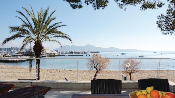 A beachside view with a dining area, a palm tree, and a marina in the background.