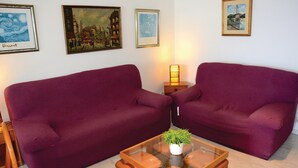 A room with two purple sofas, a glass coffee table with a plant, and three framed artworks on the wall.