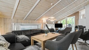 Ett mysigt vardagsrum med ett träpanelat tak, en svart lädersoffa, ett träbord och grå stolar. En platt-TV och en öppen spis syns också.