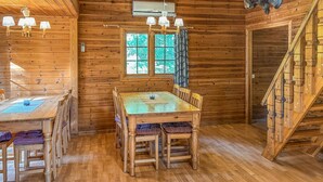 Interieur van een houten chalet met een eettafel, stoelen, een kroonluchter, een raam met gordijnen en een wenteltrap.