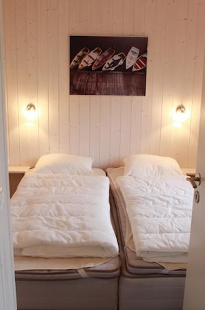 Two single beds with white mattresses and matching pillows, positioned against a wall with a wooden panel design, and a framed photograph of boats above.