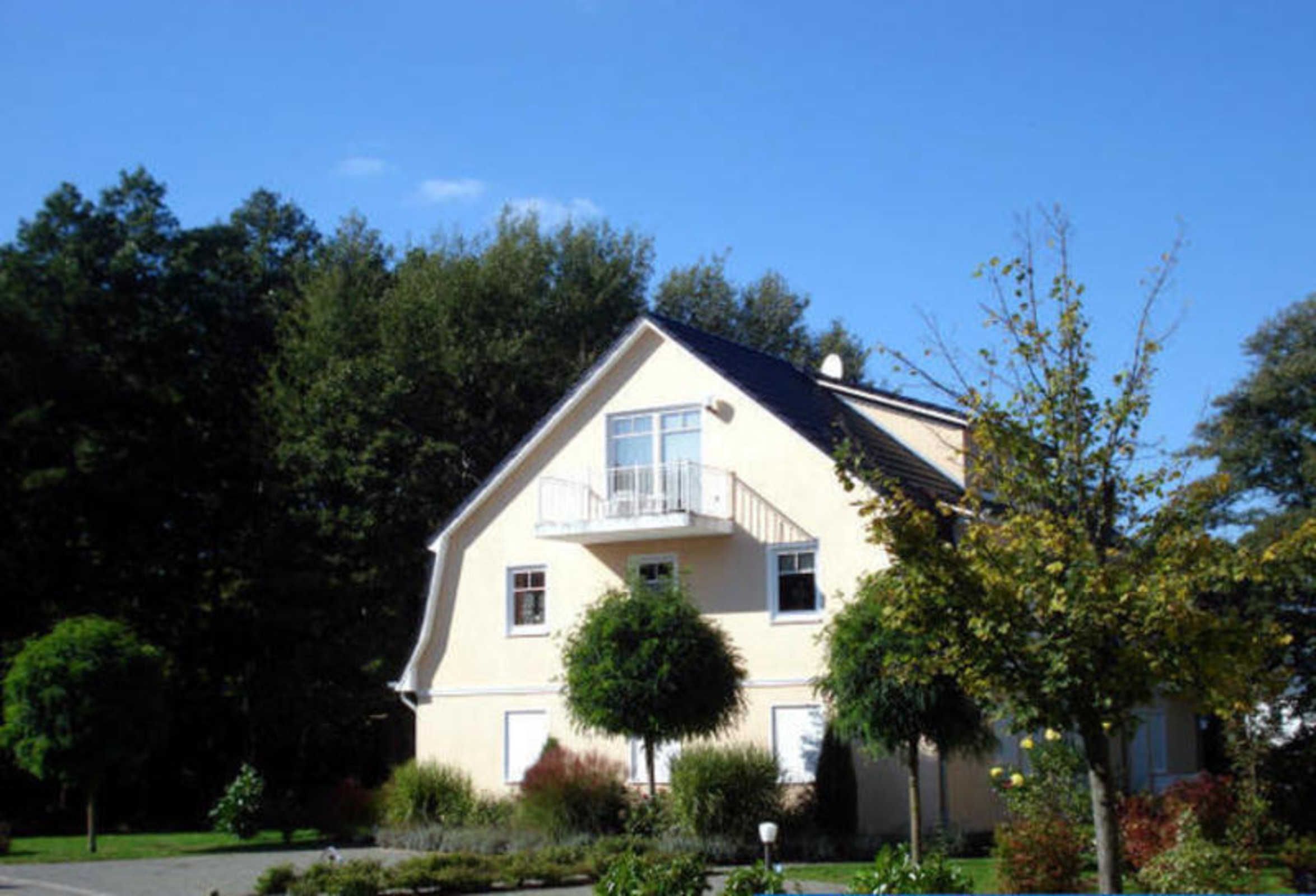 Une maison à deux étages avec un balcon, entourée d’arbres et d’un jardin bien entretenu.