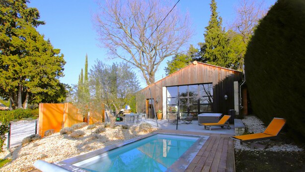 A modern outdoor swimming pool with wooden decking, a lounge area with sun loungers, and a large glass door entrance to a building. - Nice home in La Batie Rolland (La Batie Rolland)