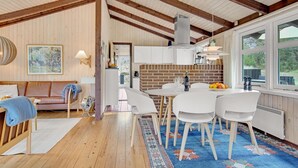 Amplia sala de estar con techo de madera, chimenea de ladrillo y comedor con sillas blancas y una mesa.