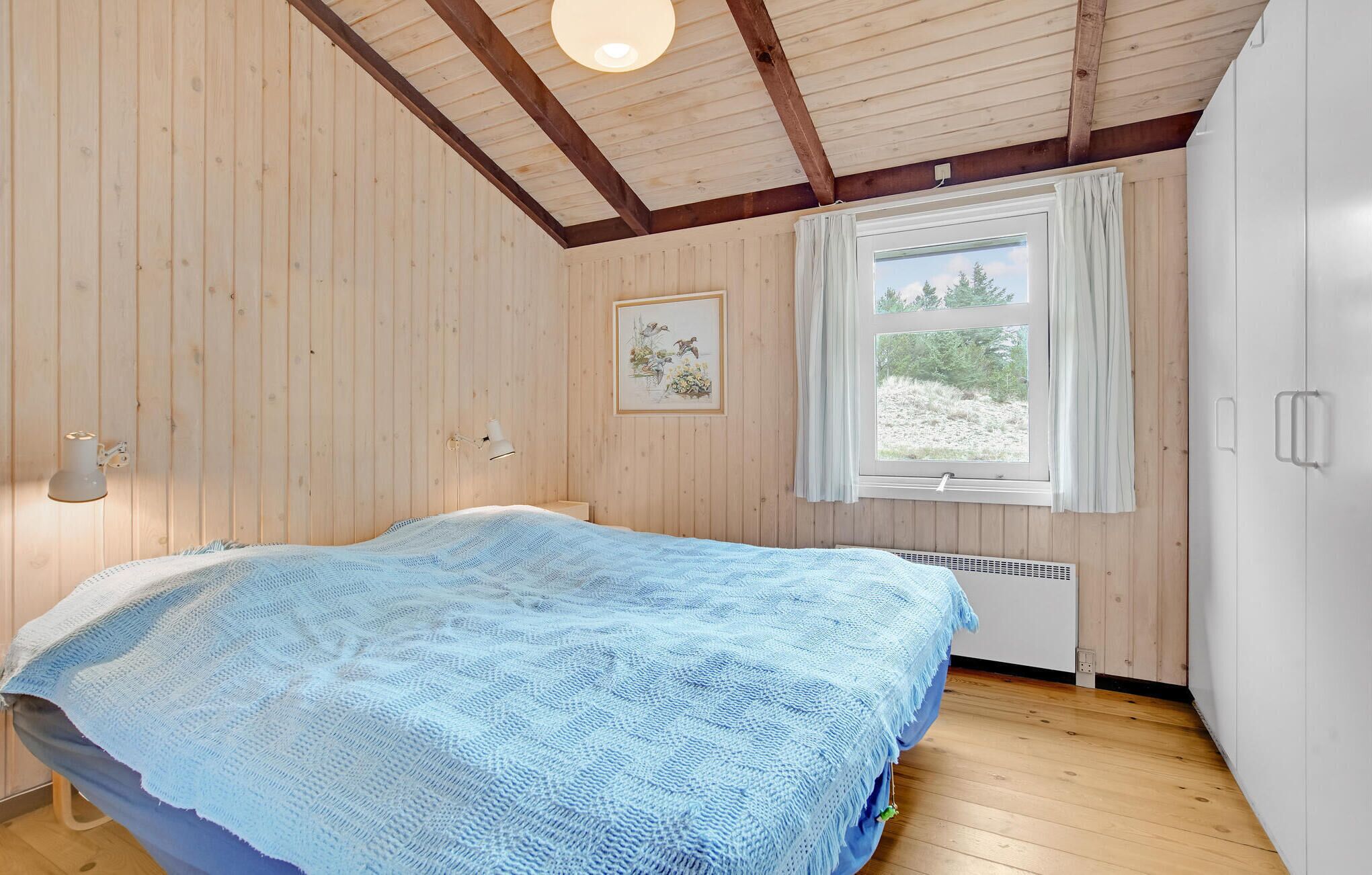 A bedroom with wooden walls, a bed with a blue blanket, a window with a view of trees, and a wall-mounted lamp.