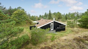 Una pequeña cabaña de madera negra con techo inclinado, rodeada de un bosque denso.