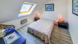 A small, well-lit room with a bed, a desk, a chair, a window, and a framed picture on the wall.