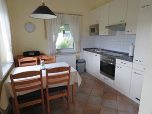 A compact kitchen with white cabinets, a stove, and a microwave. There's a dining table with chairs and a window with curtains.