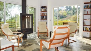 A modern living room with a fireplace, comfortable seating, and large windows offering a view of a garden and hills. - Cozy home in San Giuliano with WiFi (San Giuliano)