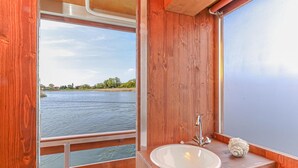 Une cabane en bois dotée d’une grande fenêtre donnant sur une rivière. À l’intérieur, on trouve un évier blanc avec un robinet moderne et un coquillage décoratif.