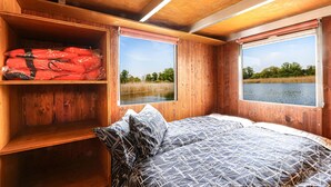 Une cabine équipée d’un lit, de deux fenêtres, d’une étagère avec des gilets de sauvetage rouges, et offrant une vue sur un lac.