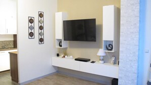 Un salon moderne avec un téléviseur à écran plat fixé au mur, un meuble de rangement blanc en dessous, et un coin cuisine sur la gauche.