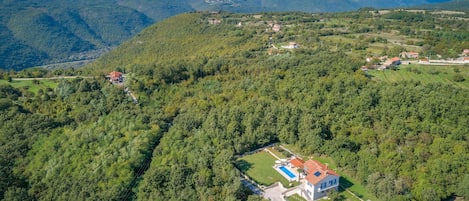 An aerial view of a property with a swimming pool, a large house, and surrounding forested hills.