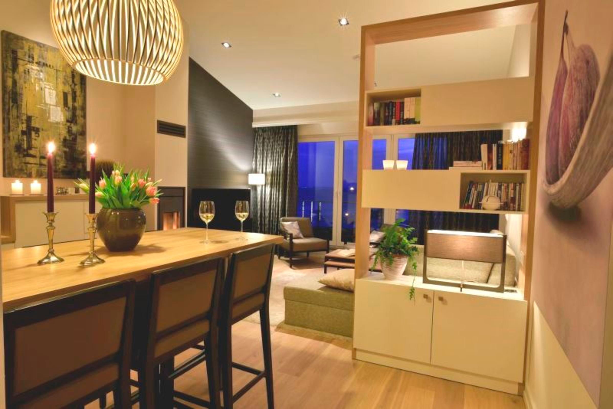 A modern living room with a wooden dining table, chairs, a bookshelf, and a large window with curtains.