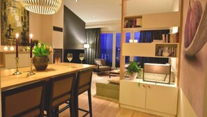 A modern living room with a wooden dining table, chairs, a bookshelf, and a large window with curtains. - Refugium Strandallee - Refugium Strandallee, 91 m², 2 - 4 persons (Neustadt in Holstein)