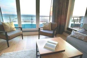 Un salon moderne doté d’une grande fenêtre donnant sur un balcon avec vue sur la mer. On y trouve deux fauteuils et un canapé avec des coussins.