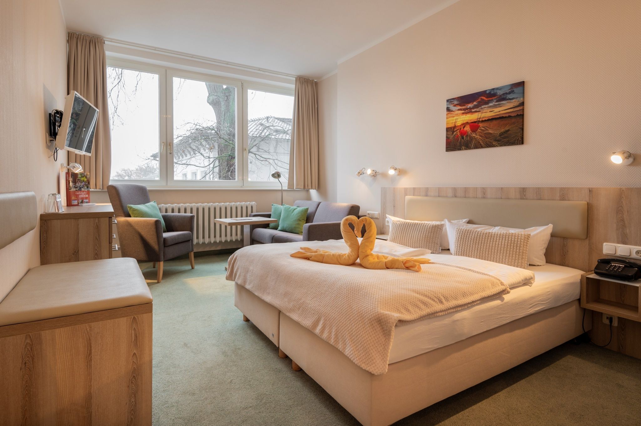 A hotel room with a bed, a desk, a chair, a radiator, a painting, and a telephone.
