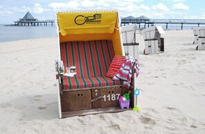 A beach chair with a yellow awning labeled 'HOTEL WALD & SEE,' a striped cushion, a small table with bottles, and a beach umbrella.