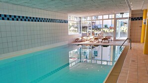 Sisätiloissa sijaitseva uima-allas, jossa on kirkasta vettä, laatoitetut seinät ja aurinkotuoleilla varustettu alue auringonottoa varten.