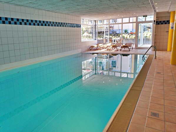 An indoor swimming pool with clear water, tiled walls, and a sunbathing area with chairs.