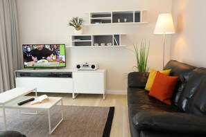 A modern living room with a flat-screen TV, a white entertainment unit, a black leather sofa with orange and yellow cushions, a white coffee table, and a floor lamp.