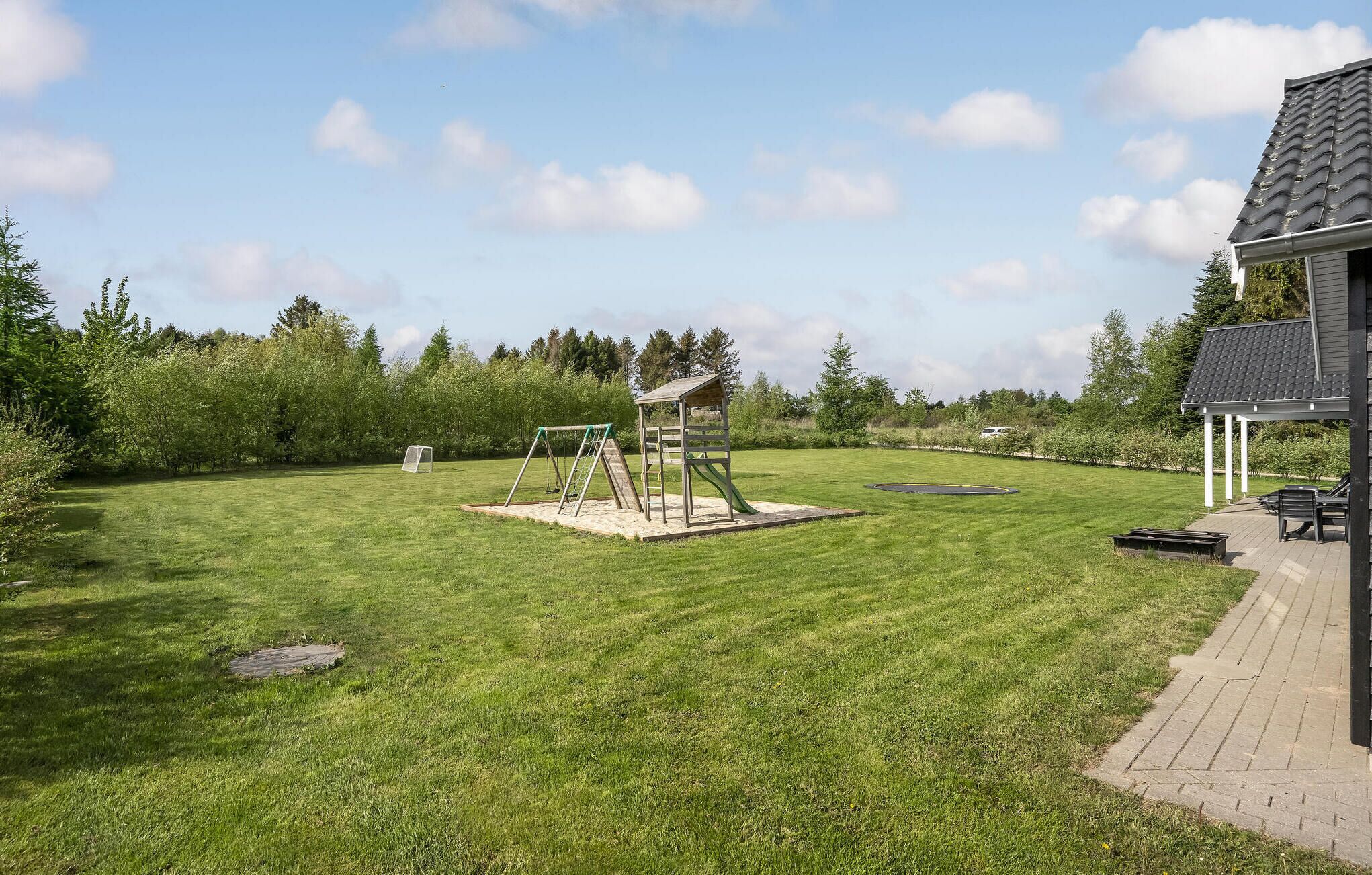 Un jardin bien entretenu, avec une balançoire, une petite maison de jeu et une terrasse dallée aménagée avec du mobilier d’extérieur.