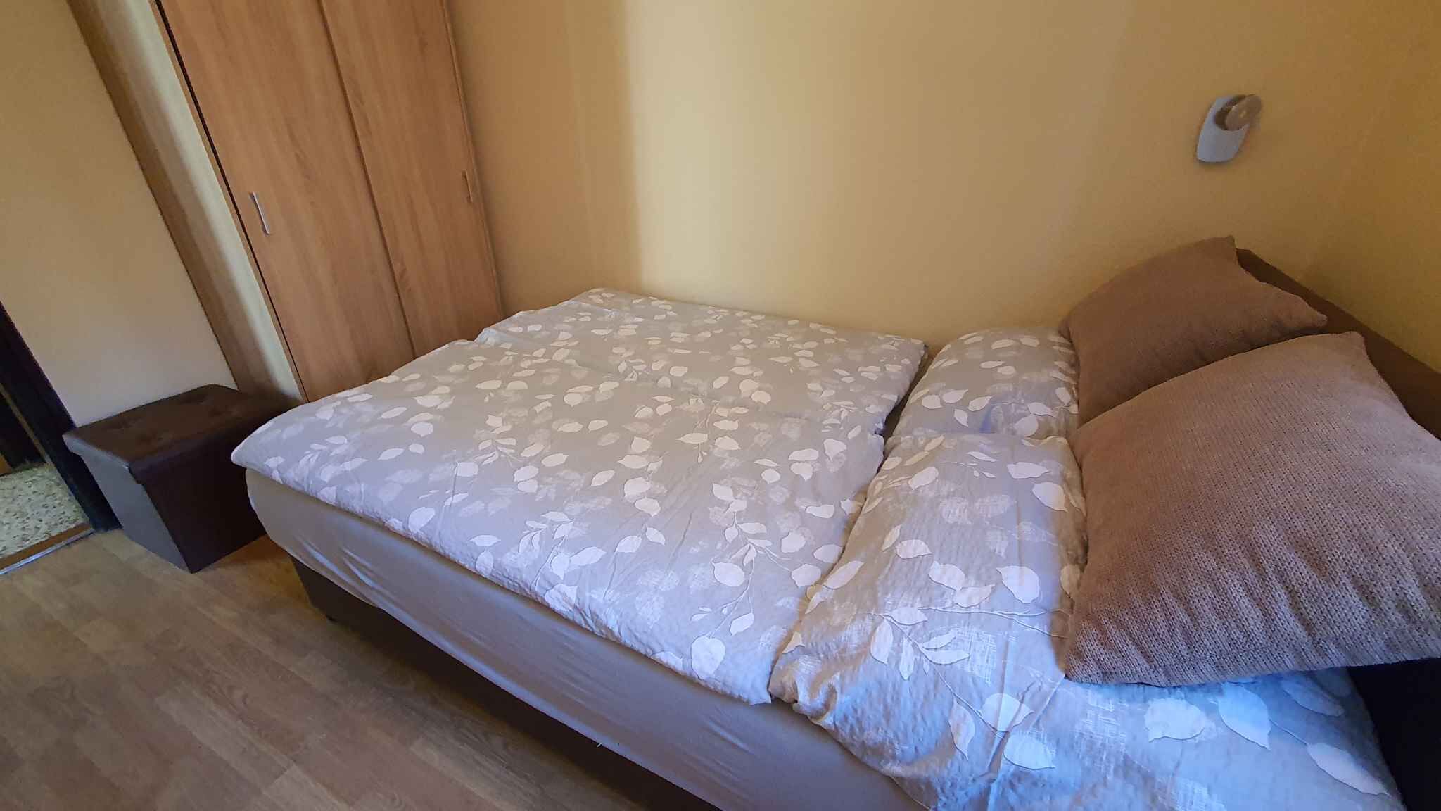 Een enkel bed met een bloemenprint sprei en twee kussens, een houten vloer en een klein nachtkastje.