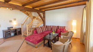 Un salon chaleureux avec un plafond en bois, un canapé d’angle rouge, une table à manger, des chaises en osier, un escalier et un meuble avec une lampe.