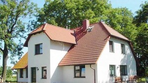 Une maison à deux étages, avec un toit recouvert de tuiles rouges et des murs blancs, entourée d’une clôture en bois et de verdure.