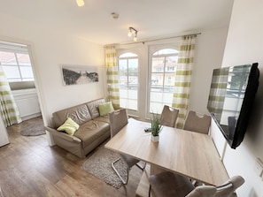 A modern living room with a wooden dining table, beige sofa, and a flat-screen TV mounted on the wall. - Haus Meeresblick A 3.19 Dünenwind with balcony - F: Haus Meeresblick A 3.19 (Baabe)