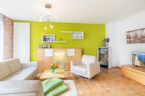 Uma sala de estar moderna com uma parede verde de destaque, um sofá, uma poltrona, uma mesa de centro com um vaso de flores, uma lareira e uma televisão.