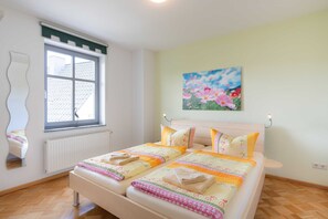 Quarto com duas camas, janela com persianas, radiador, pintura de flores e espelho na parede.