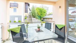 A balcony with a glass table and chairs, a small potted plant, and a view of neighboring buildings and trees.