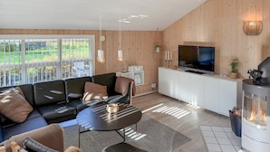 A modern living room with a black leather sofa, a round coffee table, and a TV on a white cabinet. There's a fireplace and a large window with a view of greenery. - Cozy home in Oksbøl with sauna (Oksbøl)