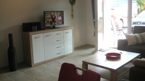 A living room with a television on a cabinet, a dining table with a red bowl, and a sofa. - 002 re (BC 3) - BUE - House Stüven (Büsum)