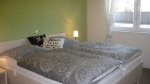 A bed with patterned bedding, pillows with text, a bedside lamp, and a window with curtains. - 002 re (BC 3) - BUE - House Stüven (Büsum)