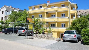 A yellow multi-story building with a sign indicating a tourist agency. There are cars parked in front, and a stone wall with plants. - Holiday apartment with balcony and air conditioning (Baška)