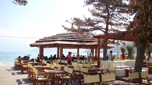 An outdoor café with wooden tables and chairs, a thatched-roof pavilion, and a view of the sea. - Holiday apartment with balcony and air conditioning (Baška)
