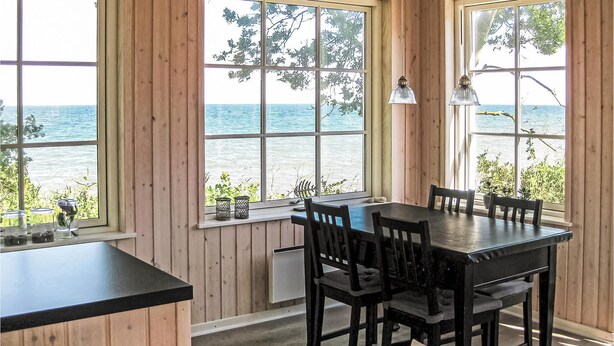 A wooden interior with a table and chairs, large windows with a view of the sea, and a lamp on the windowsill.