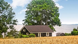 A house with a dark roof and white trim, surrounded by a field of golden wheat, with a large tree and a body of water in the background.