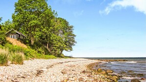 A coastal scene with a small house on a grassy hill, a pebble beach, and the ocean in the background.