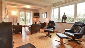 Een ruime woonkamer met houten vloeren, leren banken, een salontafel en een televisie. Grote ramen bieden natuurlijk licht en uitzicht op de omgeving.