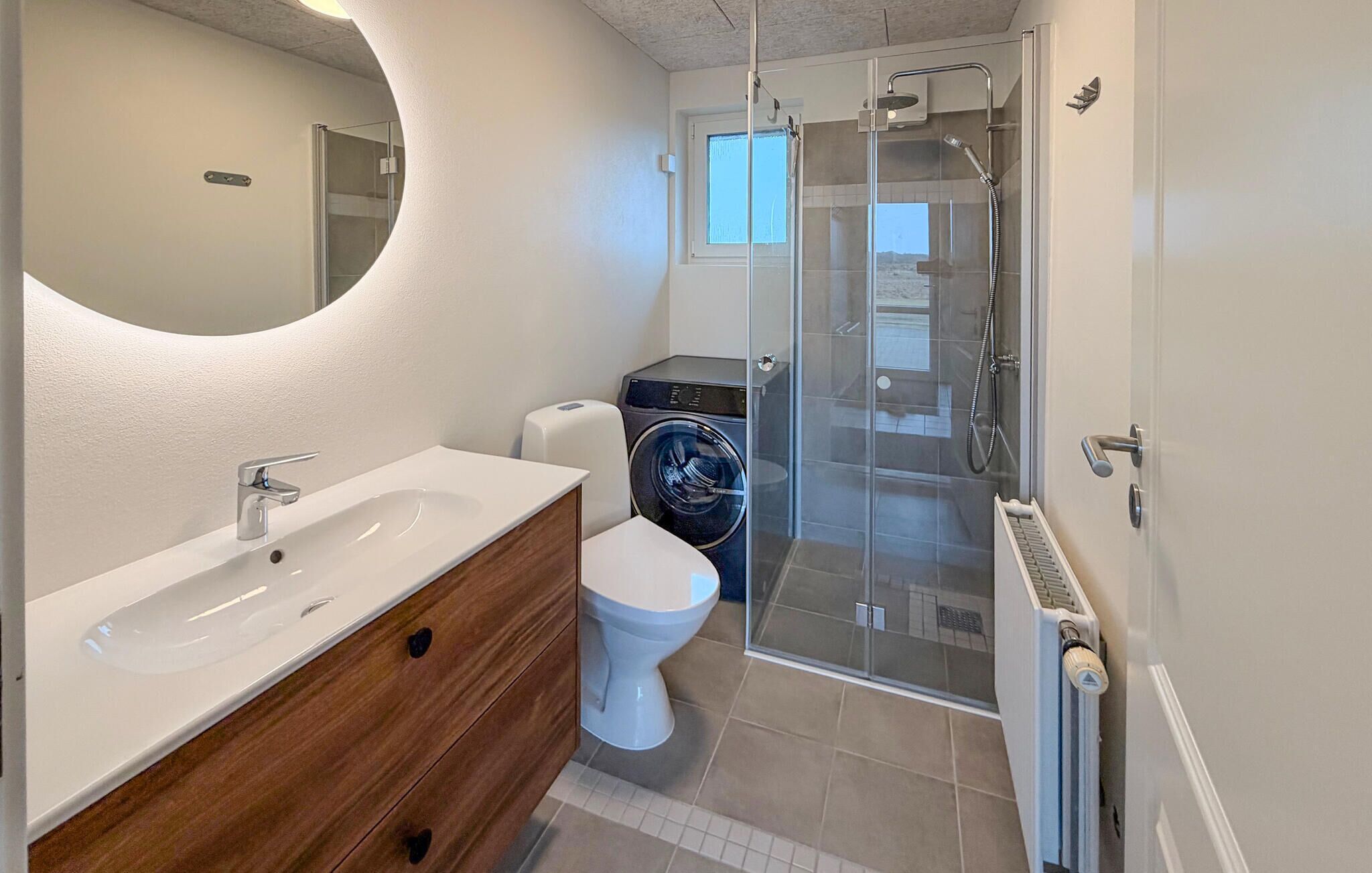 A modern bathroom with a white sink, wooden vanity, white toilet, glass shower enclosure, and a washing machine.
