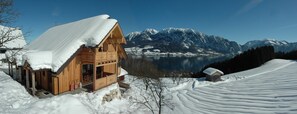 A wooden cabin with a snow-covered roof, a wooden porch, and a view of a lake and mountains in the background.