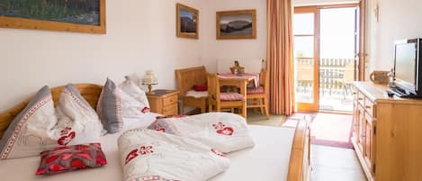 A bedroom with a bed, a wooden desk, a chair, a television, and a balcony with a view of the mountains.